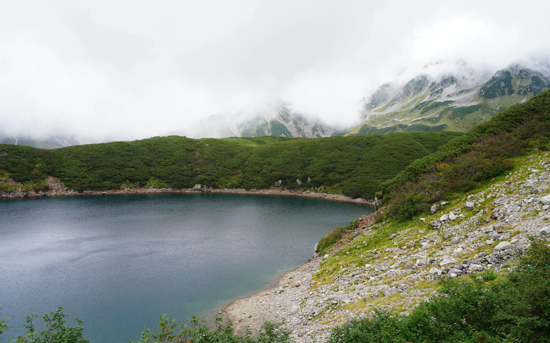 A still blue-green alpine lake beneath green hills and low cloud